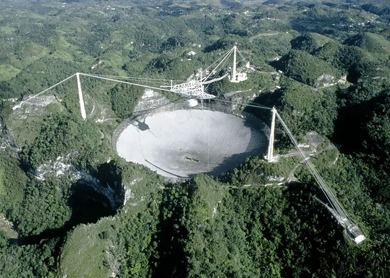 Arecibo radio observatory. Aerial view of the Arecibo radio telescope, with its gregorian subreflector system (centre). This radio telescope is situated in a natural crater in the mountains of Puerto Rico. The subreflector system has two mirrors and is housed in a 90-tonne dome receiver seen suspended 130 metres above the fixed dish. To scan the sky the dome is steered above the 305 metre dish. The Arecibo radio telescope is used by the SETI (Search for Extraterrestrial Intelli- gence) institute amongst others. SETI is an organisation dedicated to investigating evidence of intelligent civilisations elsewhere in the universe.